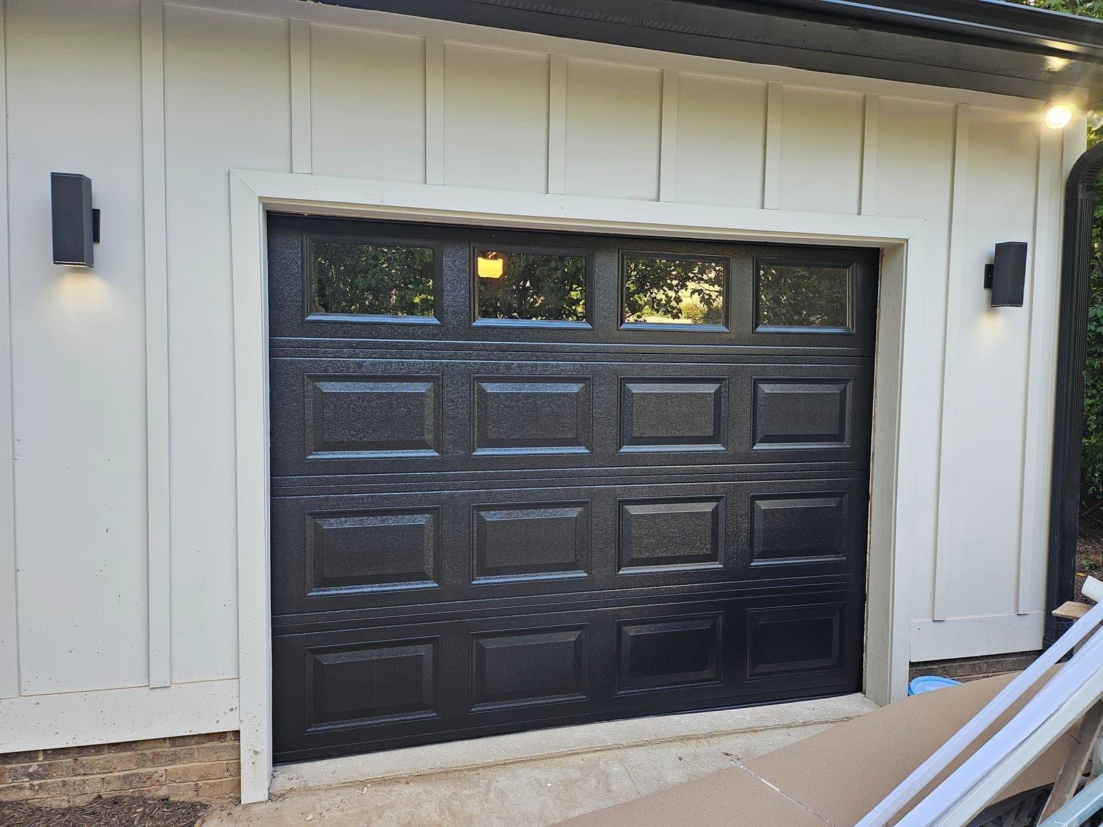 Black raised-panel garage door with window inserts on white board-and-batten home with modern sconces NC